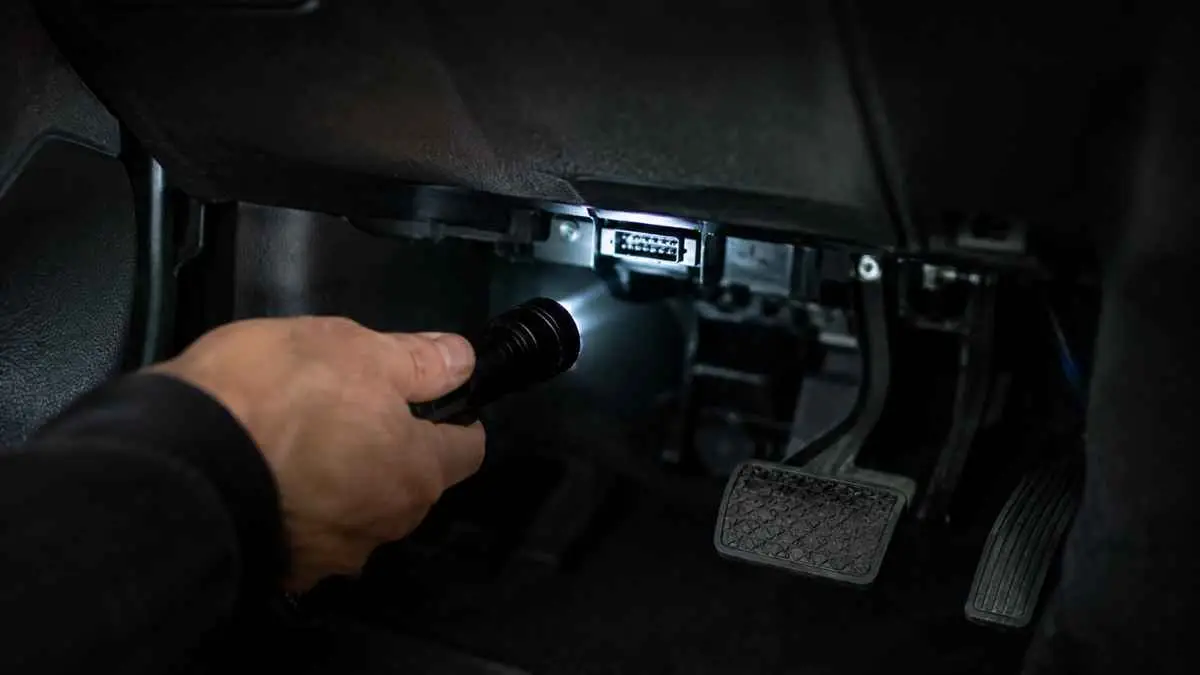 Hand with a flashlight inspecting the diagnostic port area under a car dashboard