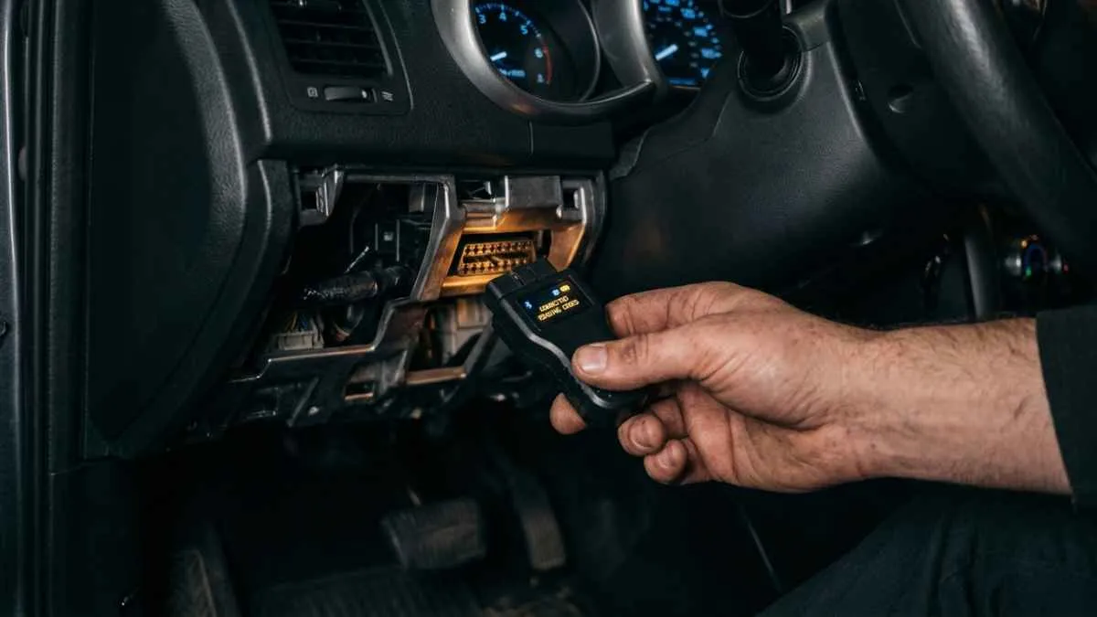 Hand holding a Bluetooth OBD2 scanner plugged into a car's diagnostic port under the dashboard