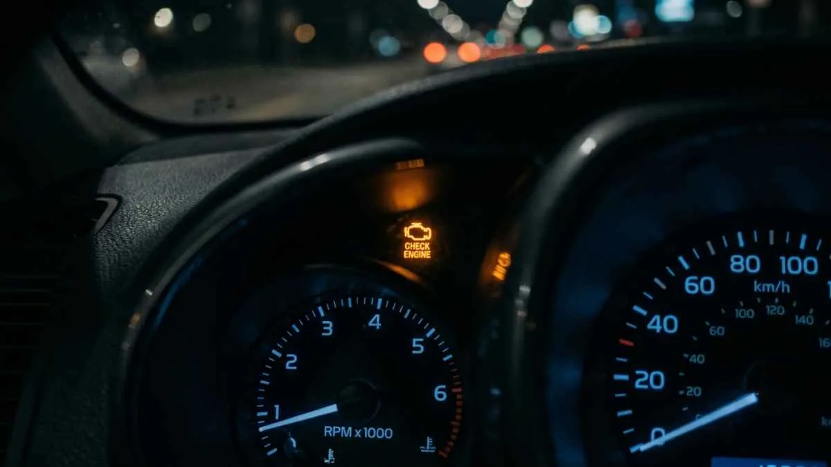 Close-up of an illuminated check engine warning light on a car dashboard at night with blurred traffic lights in the background